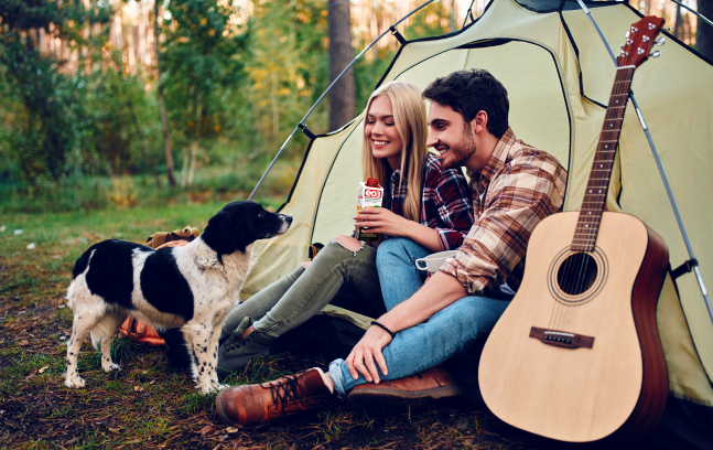 A couple sitting outside their tent with their dog and guitar in the forest. The woman is holding a Drink EO3 carton.